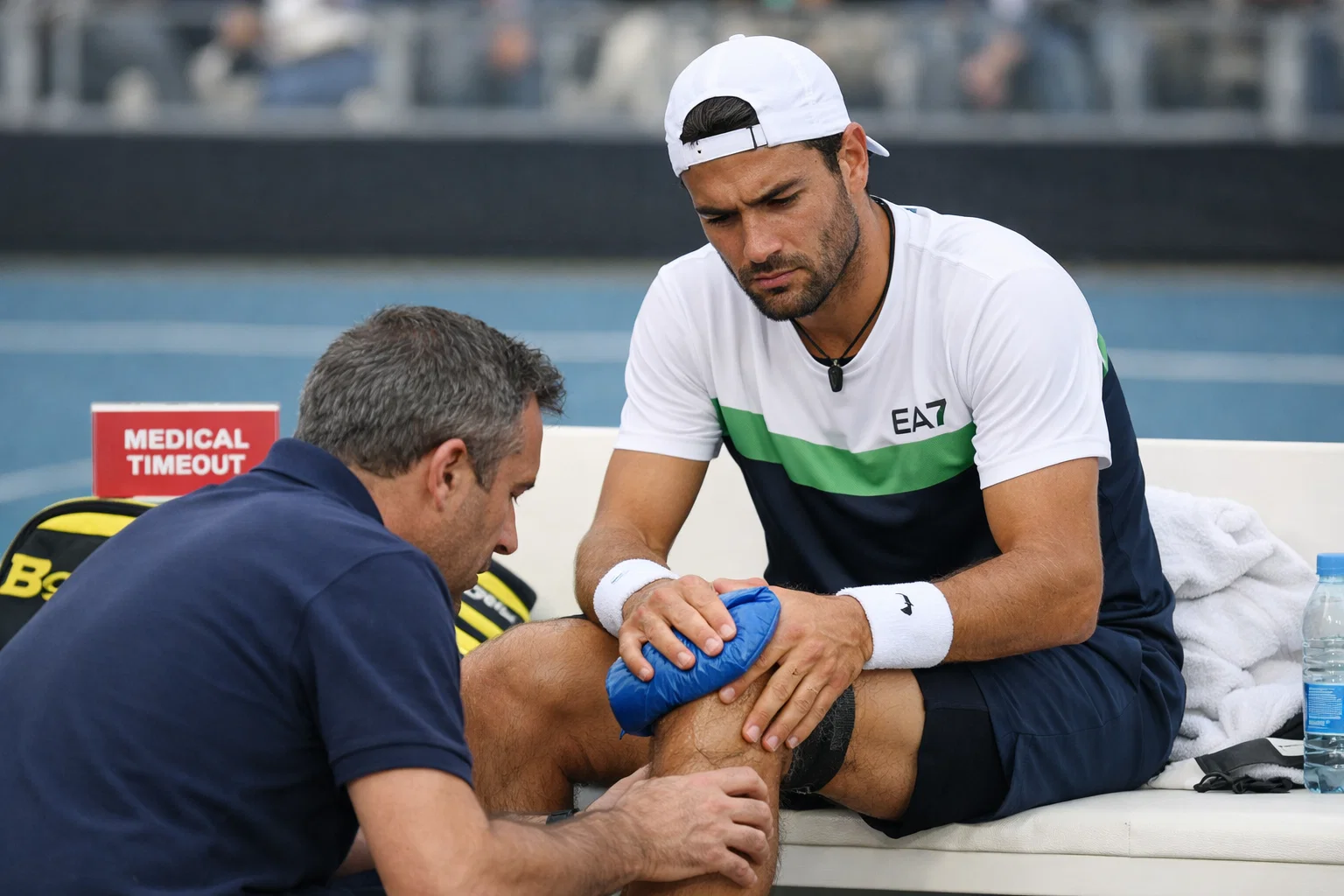 Giocatore di tennis seduto in panchina che si tiene il ginocchio con ghiaccio, fisioterapista accanto, durante una pausa medica